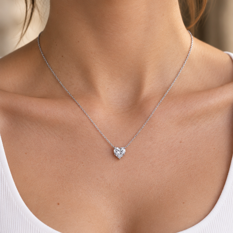 Jewelry photoshoot (worn)
Show the jewelry piece from the image worn by an elegant model in a professional editorial photograph, soft studio lighting. Reproduce the jewelry's exact design, metal, stones and details faithfully. Photorealistic, no text. Additional instructions: "Close-up editorial jewelry photo of a woman's neck, collarbone, and upper chest, cropped so the face is fully out of frame (only neck, collarbone, and chest visible). She wears a plain white top and a delicate sterling silver necklace with a clear heart-shaped cubic zirconia pendant, set in a simple four-prong setting.
METAL FINISH: the sterling silver must be rhodium-plated — bright, cool-white, and reflective with a polished mirror-like shine, not dull or matte. The metal should catch light naturally, without looking overly glossy, plastic, or artificially glowing.
STONE DETAIL: the clear heart-cut cubic zirconia stone must be sparkling and perfectly faceted with a clean heart shape, catching light realistically with natural brilliance — not flat or dull.
SKIN REALISM REQUIREMENT: skin must look completely natural and photorealistic — visible fine skin texture, natural pores, subtle imperfections, soft even tone. Avoid any smooth, plastic, airbrushed, CGI, or overly retouched look. Lighting soft and warm, casting natural gentle shadows.
STRICT SIZE REQUIREMENT: the pendant must be exactly 0.7cm height x 0.7cm width, true to real-life scale against the neck and collarbone — do not enlarge or oversize.
CHAIN AND PENDANT POSITION: fine cable chain draping naturally in a soft V-shape along the collarbone, with the heart pendant hanging centered at the lowest point of the chain, point of the heart facing down, attached by a small prong setting at the top.
Background softly blurred, warm and neutral, shallow depth of field. Mood: elegant, minimal, intimate — natural high-end jewelry brand photography style, matching the reference close-up shot, face not shown..