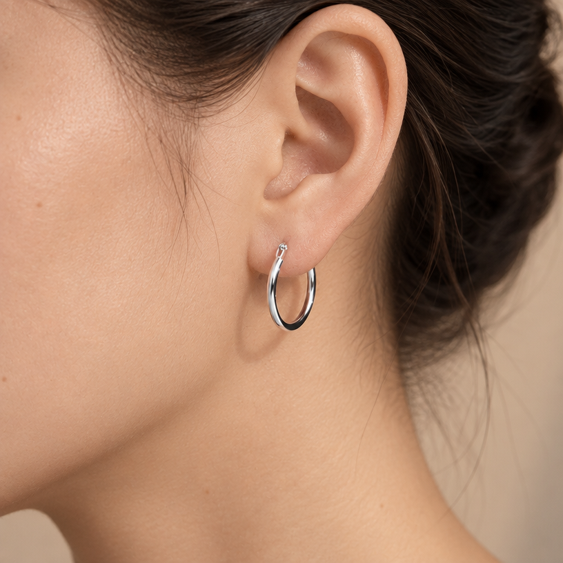 Jewelry photoshoot (worn)
Show the jewelry piece from the image worn by an elegant model in a professional editorial photograph, soft studio lighting. Reproduce the jewelry's exact design, metal, stones and details faithfully. Photorealistic, no text. Additional instructions: Close-up editorial jewelry photo of a woman's ear and jawline, tightly cropped so the face is fully out of frame (only ear, jaw edge, and neck visible). Hair pulled back or tucked behind the ear.
VARY THE POSE: do not repeat the same head tilt, shoulder position, or framing as previous generations. Use natural variations such as: head turned left or right, chin slightly up or down, hair swept to either side or tucked back, camera angle shifted slightly higher or lower. Keep the framing tight on the ear (face always out of frame), but make the pose feel distinct and natural.
She wears a sterling silver classic round hoop earring, smooth and polished, with a simple thin circular band and a hinged snap closure.
METAL FINISH: the sterling silver must be rhodium-plated — bright, cool-white, and reflective with a polished mirror-like shine, not dull or matte. The metal should catch light naturally with a smooth, glossy highlight along the curved band, without looking overly glossy, plastic, or artificially glowing.
STRICT SIZE REQUIREMENT: the hoop earring must be exactly 2cm diameter x 0.2cm band thickness, matching real-life scale against the ear — do not enlarge or oversize. The hoop should look thin, light, and delicate, not chunky or bulky.
SKIN REALISM REQUIREMENT: skin must look completely natural and photorealistic — visible fine skin texture, natural pores, subtle imperfections, soft even tone. Avoid any smooth, plastic, airbrushed, CGI, or overly retouched look. Lighting soft and warm, casting natural gentle shadows.
EARRING POSITION: the hoop sits naturally through the earlobe, hanging in a perfect circle, fully visible and in focus, catching soft light along its polished curved surface.
Background softly blurred, warm and neutral, shallow depth of field. Mood: elegant, minimal, intimate — natural high-end jewelry brand photography style, face not shown, jewelry clearly and accurately rendered in true detail and correct scale.".