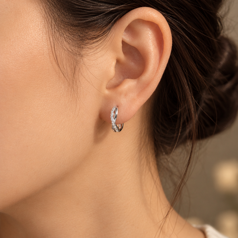 Jewelry photoshoot (worn)
Show the jewelry piece from the image worn by an elegant model in a professional editorial photograph, soft studio lighting. Reproduce the jewelry's exact design, metal, stones and details faithfully. Photorealistic, no text. Additional instructions: "Close-up editorial jewelry photo of a woman's ear and jawline, tightly cropped so the face is fully out of frame (only ear, jaw edge, and neck visible). Hair pulled back or tucked behind the ear.
VARY THE POSE: do not repeat the same head tilt, shoulder position, or framing as previous generations. Use natural variations such as: head turned left or right, chin slightly up or down, hair swept to either side or tucked back, camera angle shifted slightly higher or lower. Keep the framing tight on the ear (face always out of frame), but make the pose feel distinct and natural.
She wears a sterling silver twisted infinity hoop earring, featuring an interwoven double-band design where one smooth polished band crosses over a second band fully lined with sparkling pavé cubic zirconia stones, forming a twisted braid-like hoop.
METAL FINISH: the sterling silver must be rhodium-plated — bright, cool-white, and reflective with a polished mirror-like shine, not dull or matte. The metal should catch light naturally and sparkle realistically, without looking overly glossy, plastic, or artificially glowing.
STONE DETAIL: the pavé cubic zirconia stones must be clear, sparkling, and perfectly set in a continuous line along the twisted band, catching light realistically with natural brilliance.
STRICT SIZE REQUIREMENT: the hoop earring must be exactly 1.4cm height x 0.3cm band width, matching real-life scale against the ear — do not enlarge or oversize.
SKIN REALISM REQUIREMENT: skin must look completely natural and photorealistic — visible fine skin texture, natural pores, subtle imperfections, soft even tone. Avoid any smooth, plastic, airbrushed, CGI, or overly retouched look. Lighting soft and warm, casting natural gentle shadows.
EARRING POSITION: the hoop sits naturally through the earlobe, hanging with the twisted pattern facing forward and clearly visible, catching soft light along the pavé band.
Background softly blurred, warm and neutral, shallow depth of field. Mood: elegant, minimal, intimate — natural high-end jewelry brand photography style, face not shown, jewelry clearly and accurately rendered in true detail and correct scale..