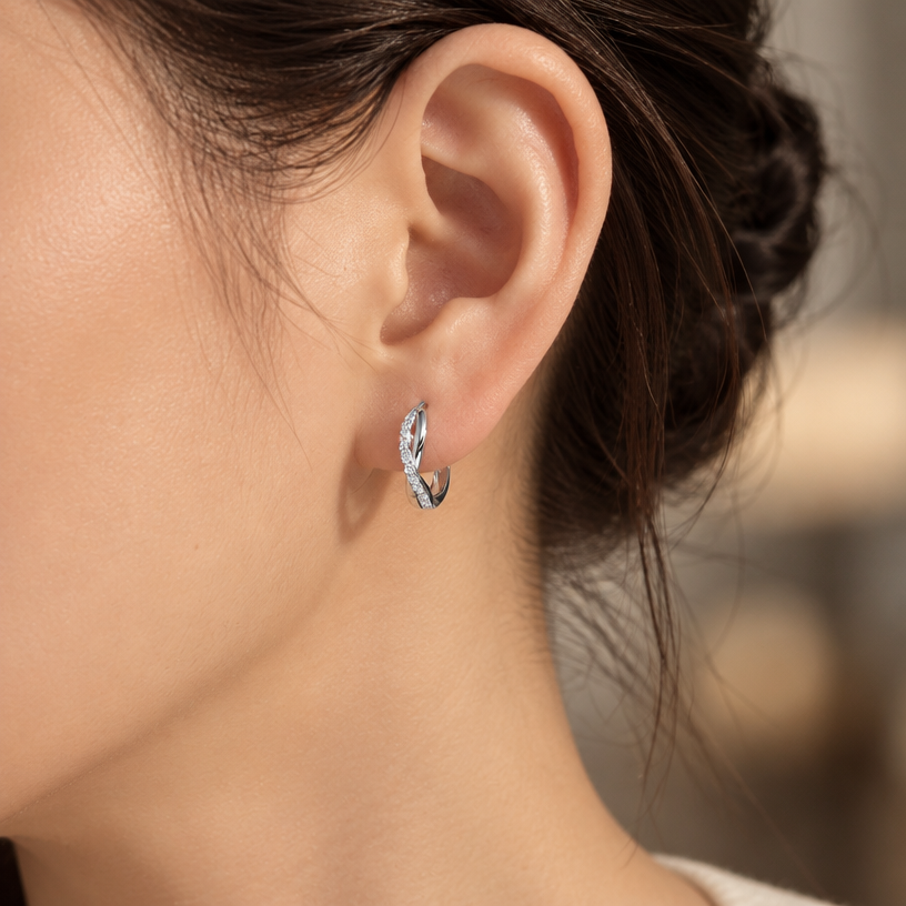 Jewelry photoshoot (worn)
Show the jewelry piece from the image worn by an elegant model in a professional editorial photograph, soft studio lighting. Reproduce the jewelry's exact design, metal, stones and details faithfully. Photorealistic, no text. Additional instructions: "Close-up editorial jewelry photo of a woman's ear and jawline, tightly cropped so the face is fully out of frame (only ear, jaw edge, and neck visible). Hair pulled back or tucked behind the ear.
VARY THE POSE: do not repeat the same head tilt, shoulder position, or framing as previous generations. Use natural variations such as: head turned left or right, chin slightly up or down, hair swept to either side or tucked back, camera angle shifted slightly higher or lower. Keep the framing tight on the ear (face always out of frame), but make the pose feel distinct and natural.
She wears a sterling silver twisted infinity hoop earring, featuring an interwoven double-band design where one smooth polished band crosses over a second band fully lined with sparkling pavé cubic zirconia stones, forming a twisted braid-like hoop.
METAL FINISH: the sterling silver must be rhodium-plated — bright, cool-white, and reflective with a polished mirror-like shine, not dull or matte. The metal should catch light naturally and sparkle realistically, without looking overly glossy, plastic, or artificially glowing.
STONE DETAIL: the pavé cubic zirconia stones must be clear, sparkling, and perfectly set in a continuous line along the twisted band, catching light realistically with natural brilliance.
STRICT SIZE REQUIREMENT: the hoop earring must be exactly 1.4cm height x 0.3cm band width, matching real-life scale against the ear — do not enlarge or oversize.
SKIN REALISM REQUIREMENT: skin must look completely natural and photorealistic — visible fine skin texture, natural pores, subtle imperfections, soft even tone. Avoid any smooth, plastic, airbrushed, CGI, or overly retouched look. Lighting soft and warm, casting natural gentle shadows.
EARRING POSITION: the hoop sits naturally through the earlobe, hanging with the twisted pattern facing forward and clearly visible, catching soft light along the pavé band.
Background softly blurred, warm and neutral, shallow depth of field. Mood: elegant, minimal, intimate — natural high-end jewelry brand photography style, face not shown, jewelry clearly and accurately rendered in true detail and correct scale..