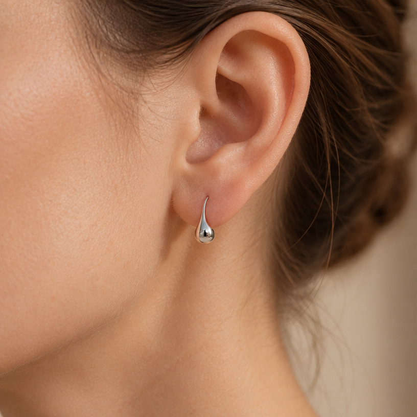 Jewelry photoshoot (worn)
Show the jewelry piece from the image worn by an elegant model in a professional editorial photograph, soft studio lighting. Reproduce the jewelry's exact design, metal, stones and details faithfully. Photorealistic, no text. Additional instructions: Close-up editorial jewelry photo of a woman's ear and jawline, tightly cropped so the face is fully out of frame (only ear, jaw edge, and neck visible). Hair pulled back or tucked behind the ear.
VARY THE POSE: do not repeat the same head tilt, shoulder position, or framing as previous generations. Use natural variations such as: head turned left or right, chin slightly up or down, hair swept to either side or tucked back, camera angle shifted slightly higher or lower. Keep the framing tight on the ear (face always out of frame), but make the pose feel distinct and natural.
She wears a sterling silver teardrop-shaped drop earring on a simple French hook wire, featuring a smooth, polished, rounded teardrop pendant that tapers elegantly at the top.
METAL FINISH: the sterling silver must be rhodium-plated — bright, cool-white, and reflective with a polished mirror-like shine, not dull or matte. The metal should catch light naturally with a smooth, glossy highlight along the curved teardrop surface, without looking overly glossy, plastic, or artificially glowing.
STRICT SIZE REQUIREMENT: the earring must stay small and delicate, true to real-life scale against the ear — do not enlarge or oversize. The teardrop should look elegant and proportionate, hanging naturally from the earlobe.
SKIN REALISM REQUIREMENT: skin must look completely natural and photorealistic — visible fine skin texture, natural pores, subtle imperfections, soft even tone. Avoid any smooth, plastic, airbrushed, CGI, or overly retouched look. Lighting soft and warm, casting natural gentle shadows.
EARRING POSITION: the earring hangs naturally from the earlobe via the French hook wire, teardrop pointing straight down, fully visible and in focus, catching soft light along its curved silhouette.
Background softly blurred, warm and neutral, shallow depth of field. Mood: elegant, minimal, intimate — natural high-end jewelry brand photography style, face not shown, jewelry clearly and accurately rendered in true detail and correct scale.".