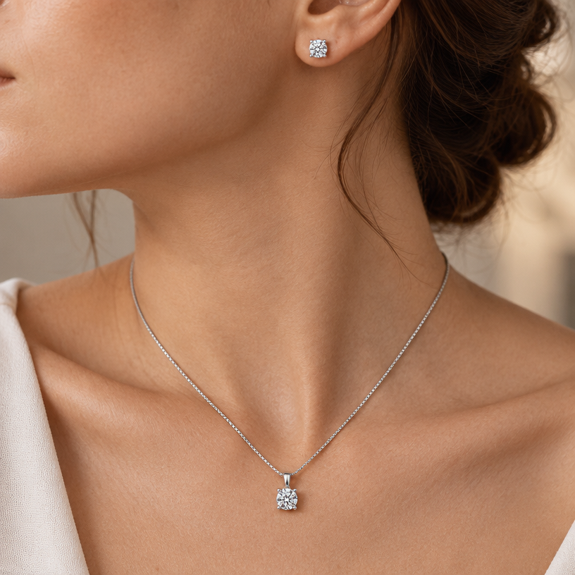 Jewelry photoshoot (worn)
Show the jewelry piece from the image worn by an elegant model in a professional editorial photograph, soft studio lighting. Reproduce the jewelry's exact design, metal, stones and details faithfully. Photorealistic, no text. Additional instructions: Close-up editorial jewelry photo of a woman's neck, collarbone, ear, and upper chest, cropped so the face is fully out of frame. She wears soft, elegant clothing, only a small part of the fabric visible at the edge of the frame.
VARY THE POSE: do not repeat the same head tilt, shoulder position, or framing as previous generations. Use natural variations such as: head turned left or right, chin slightly up or down, one shoulder raised or relaxed, hair swept to either side or tucked back, camera angle shifted slightly higher or lower. Keep the framing tight on the neck, collarbone, and ear (face always out of frame), but make the pose feel distinct and natural.
She wears a sterling silver necklace with a single round brilliant-cut clear cubic zirconia solitaire pendant set in a classic four-prong setting, and a matching pair of round brilliant-cut cubic zirconia solitaire stud earrings in the same four-prong setting.
METAL FINISH: the sterling silver must be rhodium-plated — bright, cool-white, and reflective with a polished mirror-like shine, not dull or matte. The metal should catch light naturally, without looking overly glossy, plastic, or artificially glowing.
STONE DETAIL: the round cubic zirconia stones must be clear, sparkling, and perfectly faceted with a classic round brilliant cut, catching light realistically with strong natural brilliance and fire, like a fine diamond simulant.
STRICT SIZE REQUIREMENT: the pendant and earrings must be small and delicate, true to real-life scale against the neck and ear — a classic solitaire size, not oversized or exaggerated.
SKIN REALISM REQUIREMENT: skin must look completely natural and photorealistic — visible fine skin texture, natural pores, subtle imperfections, soft even tone. Avoid any smooth, plastic, airbrushed, CGI, or overly retouched look. Lighting soft and warm, casting natural gentle shadows.
CHAIN AND PENDANT POSITION: fine box chain draping naturally in a soft V-shape along the collarbone, with the round solitaire pendant hanging centered at the lowest point of the chain, attached by a simple bail. The stud earrings sit naturally and securely on the earlobes, fully visible and in focus.
Background softly blurred, warm and neutral, shallow depth of field. Mood: elegant, minimal, intimate — natural high-end jewelry brand photography style, face not shown, jewelry clearly and accurately rendered in true detail and correct scale.".