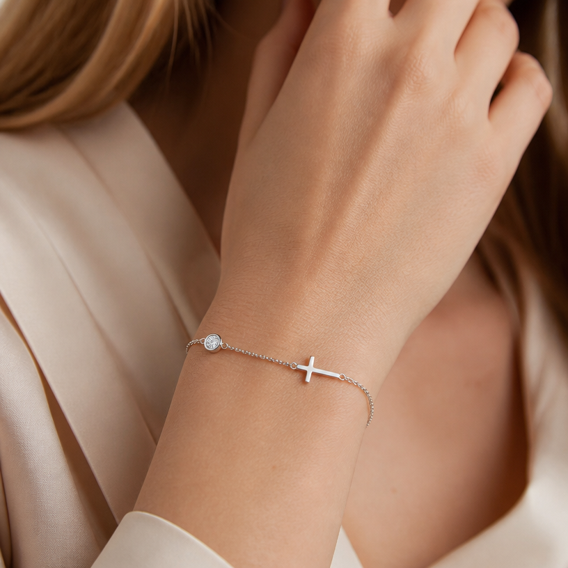 Jewelry photoshoot (worn)
Show the jewelry piece from the image worn by an elegant model in a professional editorial photograph, soft studio lighting. Reproduce the jewelry's exact design, metal, stones and details faithfully. Photorealistic, no text. Additional instructions: "Close-up beauty/editorial photo of a woman's wrist and hand, fair light skin tone, gently relaxed pose (resting on a soft surface or lightly touching her face/collarbone). Soft, warm neutral studio background (beige/cream tones), softly blurred, shallow depth of field.
She wears a delicate sterling silver chain bracelet featuring a small round bezel-set cubic zirconia stone on one side of the chain and a small sideways cross charm on the other side, connected by a fine cable chain.
METAL FINISH: the sterling silver must be rhodium-plated — bright, cool-white, and reflective with a polished mirror-like shine, not dull or matte. The metal should catch light naturally and sparkle realistically, without looking overly glossy, plastic, or artificially glowing.
SKIN REALISM REQUIREMENT: skin must look completely natural and photorealistic — fair, light skin tone to enhance the contrast and shine of the silver, with visible fine skin texture, natural pores, subtle imperfections. Avoid any smooth, plastic, airbrushed, CGI, or overly retouched look. Lighting soft and warm, casting natural gentle shadows.
STRICT SIZE REQUIREMENT: the bracelet must stay true to real-life scale, fine and delicate — the round stone approximately 0.6cm, the cross charm small and slim, both proportionate to the wrist. Do not enlarge or oversize.
BRACELET POSITION: the chain drapes naturally around the wrist, with the round stone and cross charm both visible and facing forward, sitting flat against the skin.
Mood: elegant, minimal, intimate — high-end jewelry brand photography style, sharp focus on the bracelet while keeping the composition natural and soft..