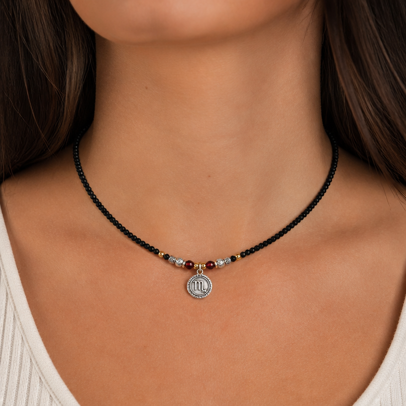 A beaded women's necklace made of mixed metal with garnet stones suitable for daily wear, detailed and decorative, moderately ornate. Simple necklace on a jewelry cord made of 3mm black tourmaline beads, approx. 40 cm long, with a small silver pendant with the scorpio zodiac sign. The background for this necklace is neck of a young woman wearing this necklace.