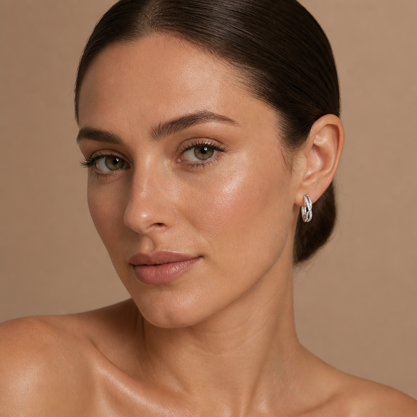 Jewelry photoshoot (worn)
Show the jewelry piece from the image worn by an elegant model in a professional editorial photograph, soft studio lighting. Reproduce the jewelry's exact design, metal, stones and details faithfully. Photorealistic, no text. Additional instructions: Front-facing beauty portrait of a woman, head slightly turned to reveal her ear and jewelry, direct calm gaze into the camera. Hair sleek and pulled back into a low bun, fully exposing the ear, neck, and shoulders. Bare shoulders, no clothing visible. Plain, smooth warm beige/nude studio background, softly lit with even, diffused lighting.
She wears a sterling silver twisted infinity hoop earring, featuring an interwoven double-band design where one smooth polished band crosses over a second band fully lined with sparkling pavé cubic zirconia stones, forming a twisted braid-like hoop.
METAL FINISH: the sterling silver must be rhodium-plated — bright, cool-white, and reflective with a polished mirror-like shine, not dull or matte. The metal should catch light naturally and sparkle realistically, without looking overly glossy, plastic, or artificially glowing.
SKIN REALISM REQUIREMENT: skin must look completely natural and photorealistic — visible fine skin texture, natural pores, subtle imperfections, soft even tone. Avoid any smooth, plastic, airbrushed, CGI, or overly retouched look. Lighting soft and flattering, casting natural gentle shadows.
STRICT SIZE REQUIREMENT: the hoop earring must be exactly 1.4cm height x 0.3cm band width, matching real-life scale against the ear — do not enlarge or oversize.
EARRING POSITION: the hoop sits naturally through the earlobe, twisted pattern facing forward and clearly visible, catching soft light along the pavé band.
Mood: elegant, high-end beauty/jewelry campaign photography, clean and polished, sharp focus on the ear and earring while keeping the face naturally in focus..